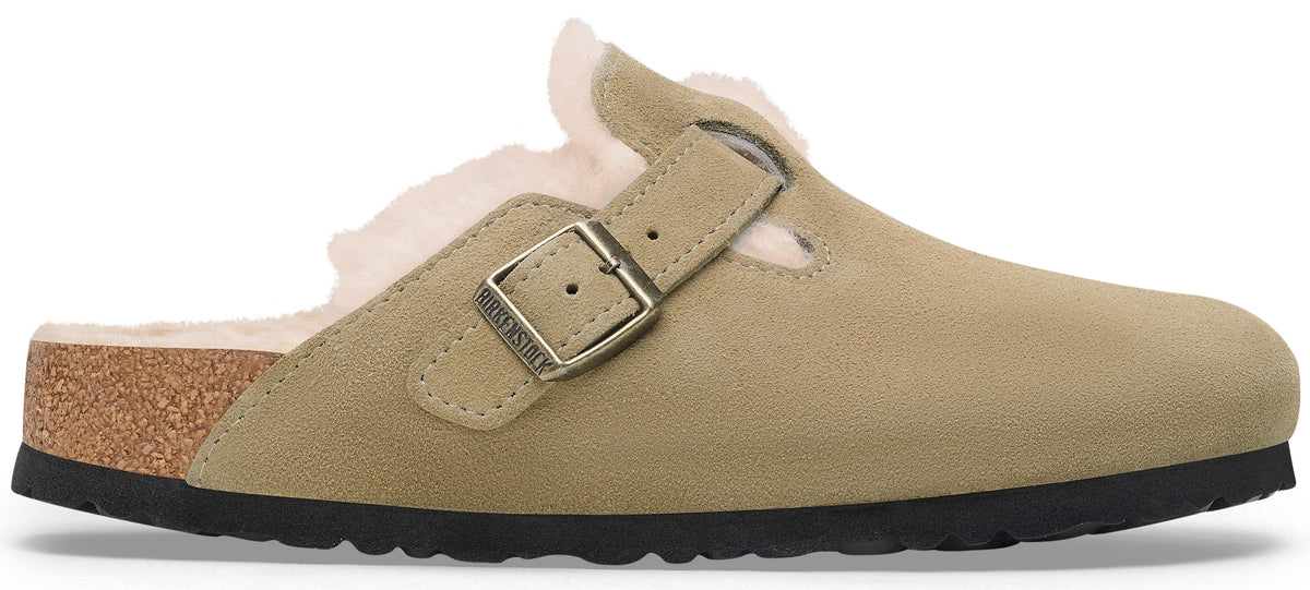A beige suede slip-on clog with a shearling lining, featuring an adjustable metal buckle strap with the embossed text 'BIRKENSTOCK', a cork midsole, and a black textured rubber outsole.