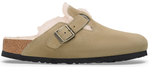 A beige suede slip-on clog with a shearling lining, featuring an adjustable metal buckle strap with the embossed text 'BIRKENSTOCK', a cork midsole, and a black textured rubber outsole.