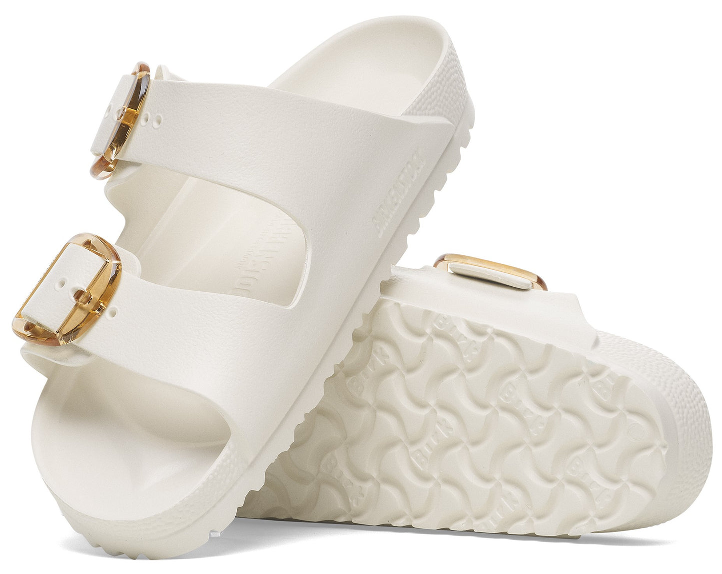 Two white slide sandals with gold buckles are angled on their sides, one behind the other.