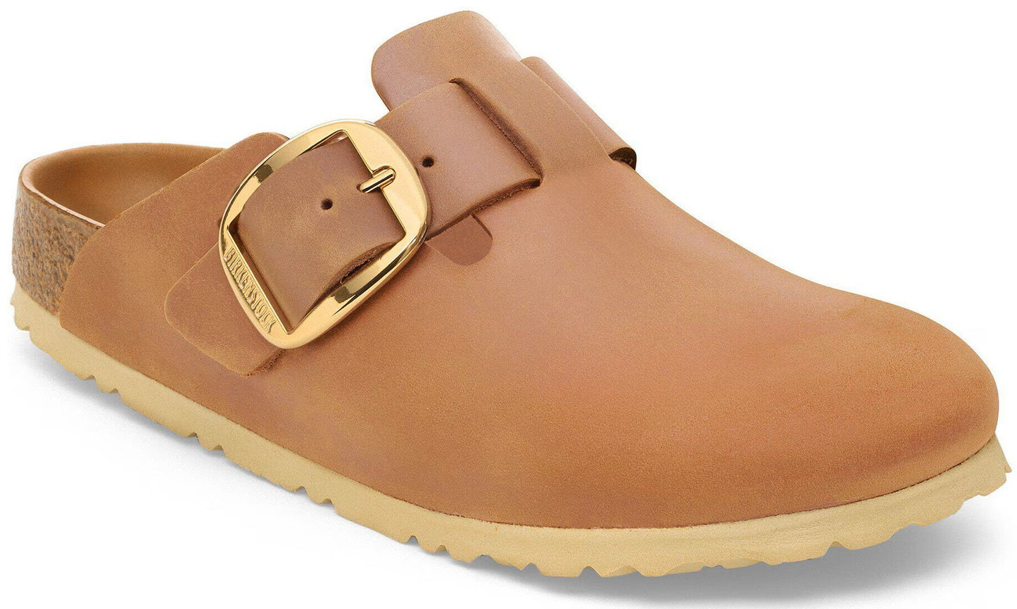 A tan leather clog shoe with an oversized polished gold metal buckle and a cork footbed liner visible at the heel, resting on a beige rubber lug sole.
