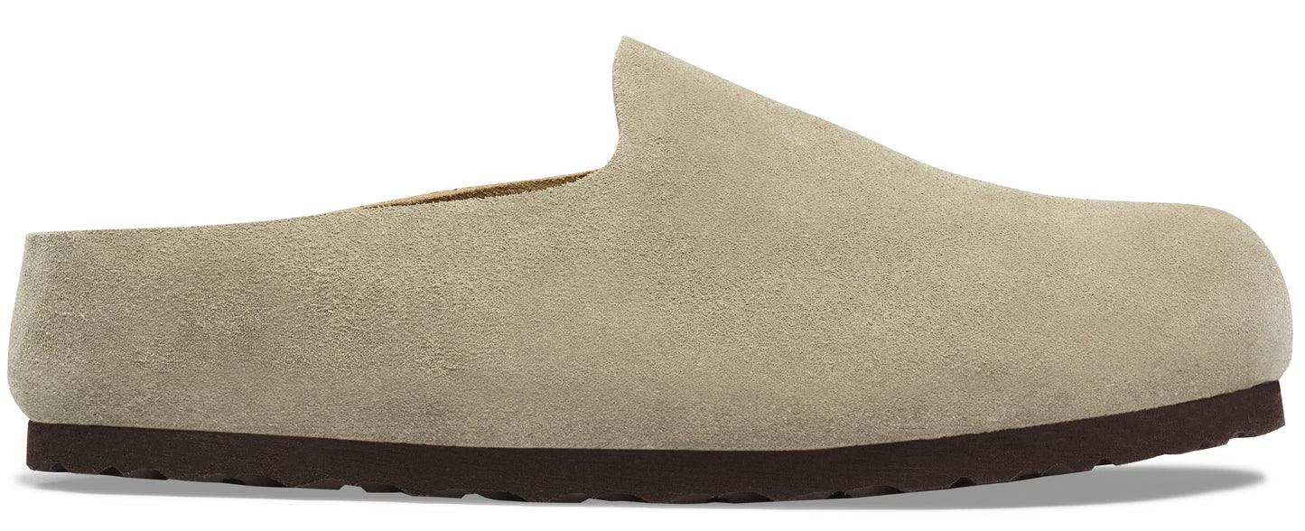A slip-on mule shoe made of tan suede with a flat, dark brown rubber sole, shown in profile.