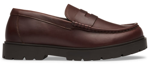 A dark brown leather penny loafer shoe featuring a classic apron toe, a strap across the instep, and a thick, lugged, dark brown rubber sole.