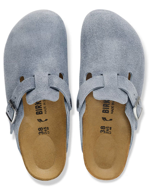 A pair of light blue suede clogs with a closed rounded toe, an adjustable buckled strap across the instep, and a cork-latex footbed with Birkenstock branding and size 38 visible on the sole.