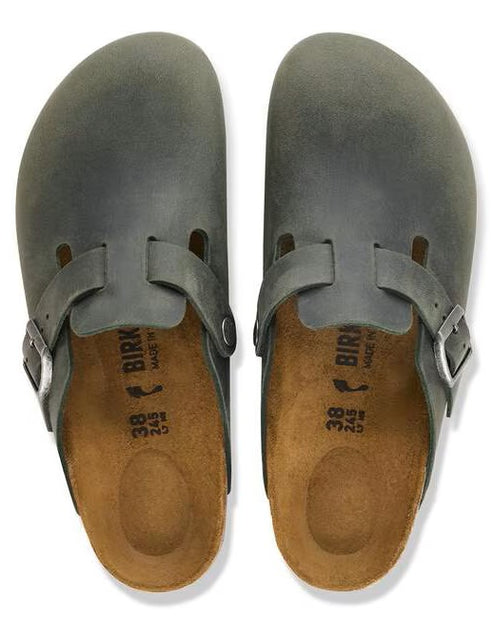 A pair of slate-grey suede leather clogs with an open back, featuring a side strap with a silver buckle, brown cork-latex footbeds with imprinted size 38, and Birkenstock brand text on a plain white background.