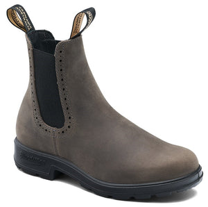 A brown leather Chelsea boot with a black elastic side panel, featuring decorative brogue-style perforations along the collar edge, two fabric pull tabs at the top, and a thick, lugged black rubber sole, with 'Blundstone' embossed on the heel.