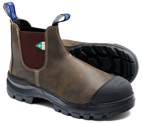 A brown leather slip-on work boot with a reinforced black rubber toe cap, brown elastic side panels, and blue fabric pull tabs on the front and back, featuring heavy-duty black lugged soles and visible stitching.