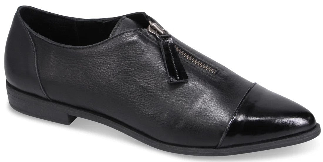 A black leather flat shoe with a pointed toe, a patent leather cap toe, and a center zipper closure with a rectangular pull tab.