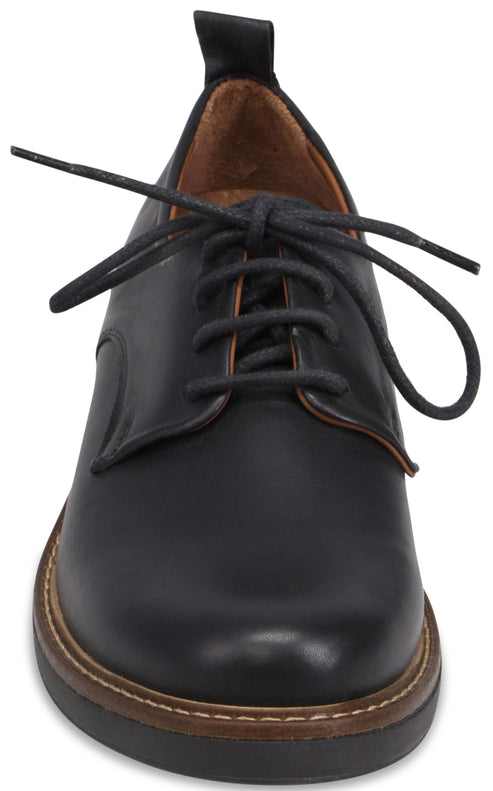 A close-up front view of a black leather derby shoe with a rounded toe, a brown welt with contrast stitching, and black laces tied in a bow.