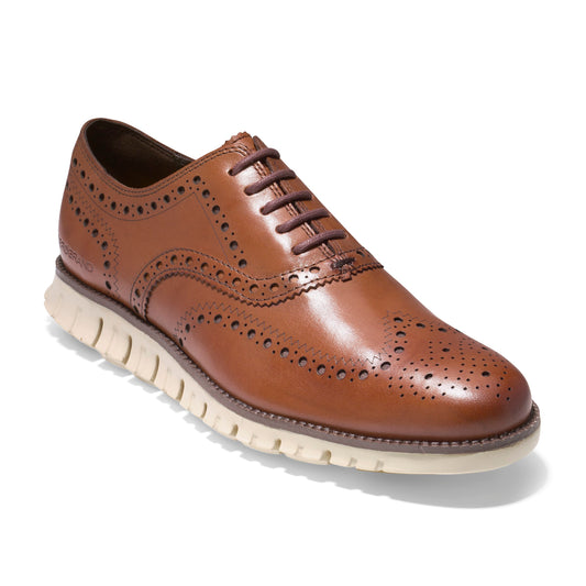 A tan leather dress shoe with decorative perforations, brown laces, and a white rubber sole.