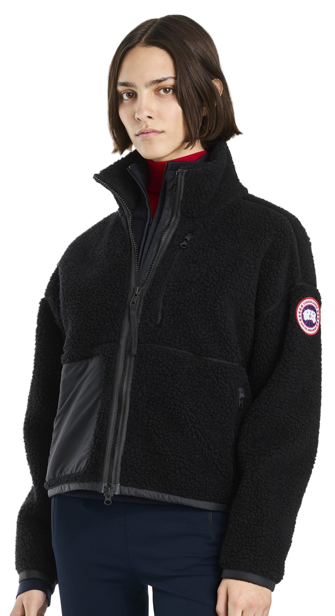 A woman is wearing a black, zip-up, fleece jacket with the Canada Goose logo on the left sleeve and a high collar.