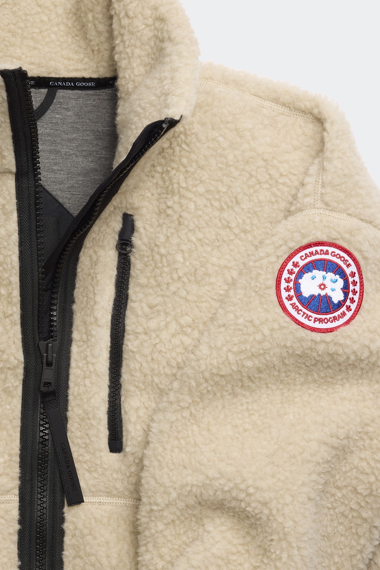 Close-up of a cream sherpa jacket with black zipper, chest pocket, and a circular patch with a maple leaf.