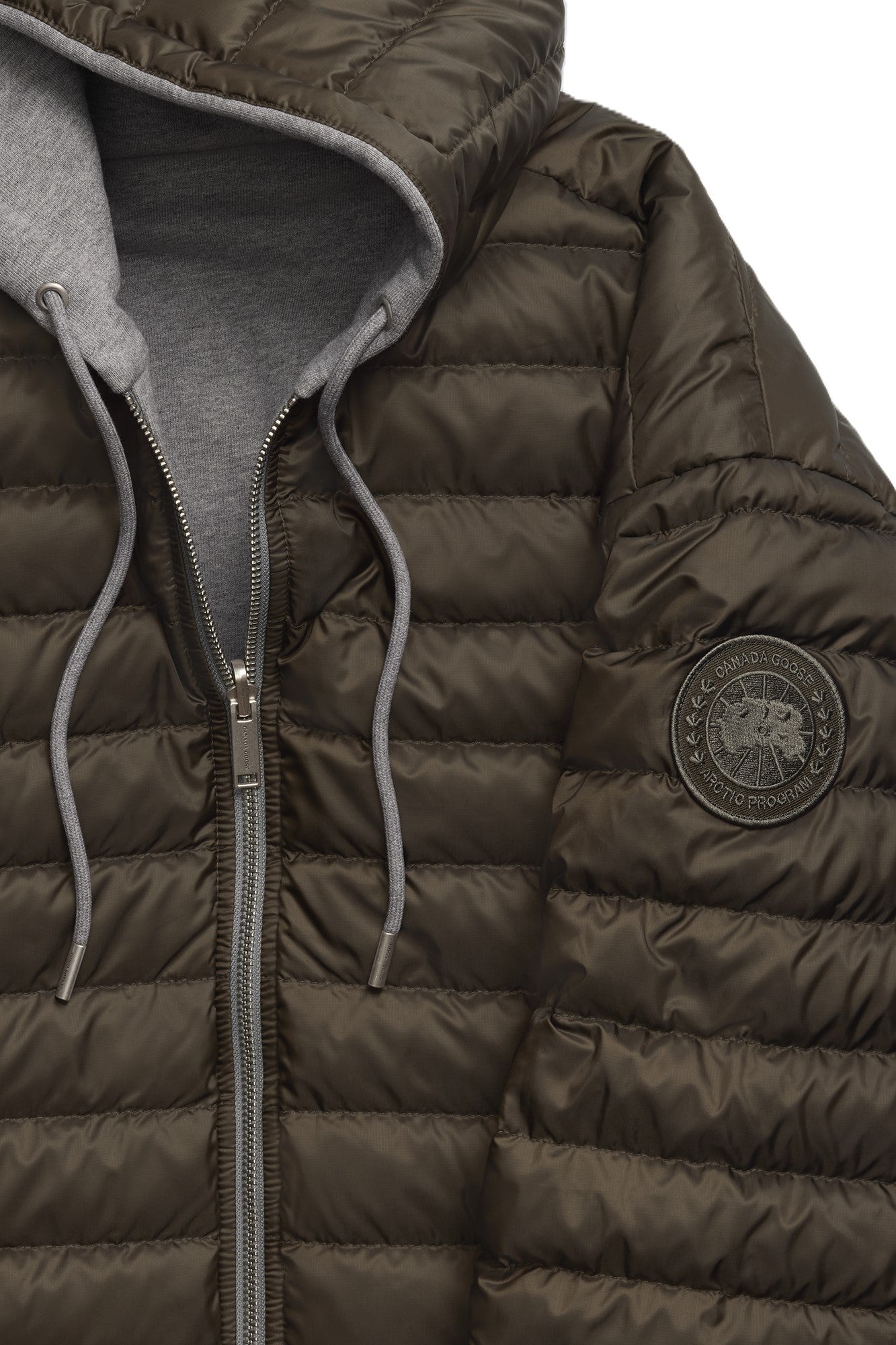 Close-up of a puffy, brown, hooded jacket with a gray hood and silver zipper. The sleeve has a round, dark-colored emblem.