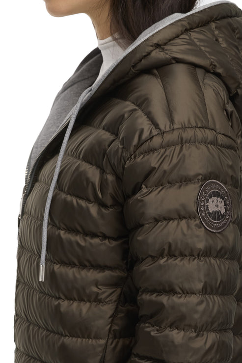 Close-up of a woman wearing an olive-green puffer jacket with a gray hood featuring a round badge on the right sleeve.