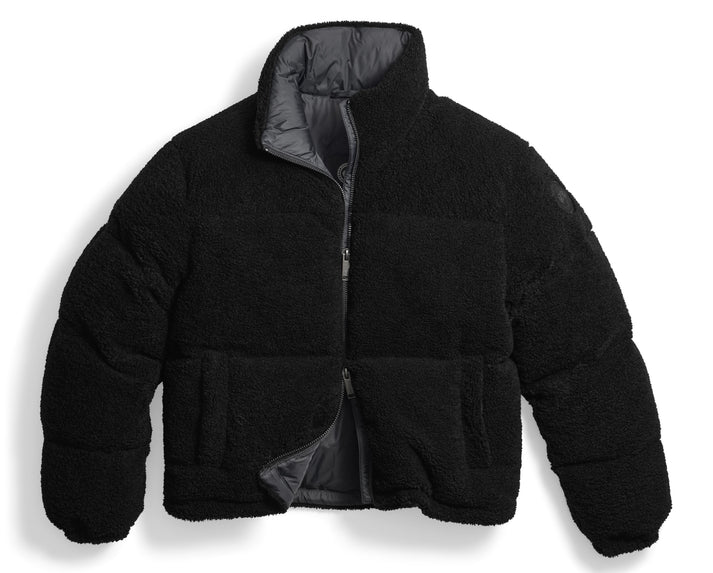 A black, cropped, puffer jacket made of textured, fleece-like material, featuring a high collar, long sleeves, and a central zipper closure that is partially open to reveal a grey interior lining.
