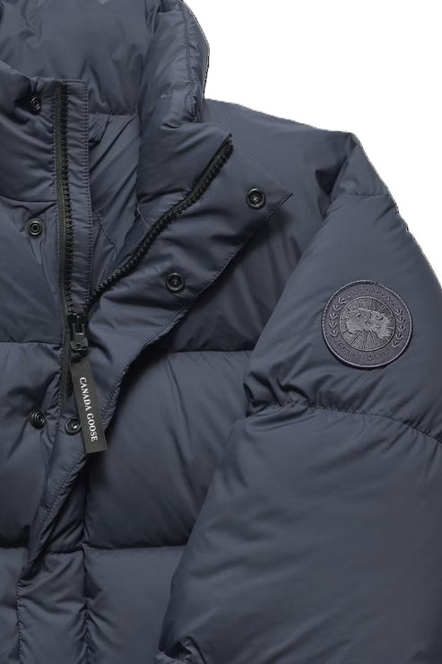A close-up view of a dark blue, puffer jacket featuring a monochromatic circular logo patch on the left arm and a black zipper pull with the words CANADA GOOSE printed in white text.