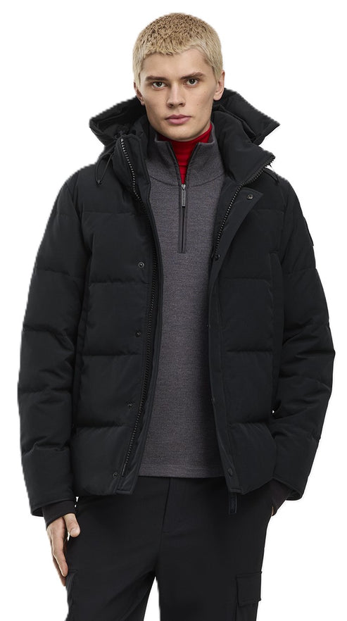 A fair-skinned man with short blonde hair wears a black hooded puffer jacket over a grey quarter-zip sweater and a red mock-neck shirt.