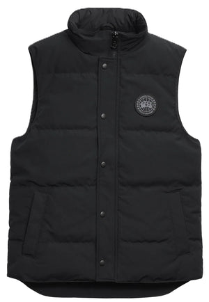 A black quilted puffer vest with a high collar, a central snap-button placket, two side pockets, and a round black and grey circular patch on the left chest.