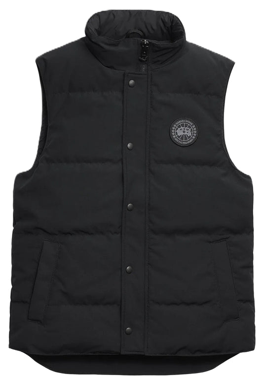 A black quilted puffer vest with a high collar, a central snap-button placket, two side pockets, and a round black and grey circular patch on the left chest.