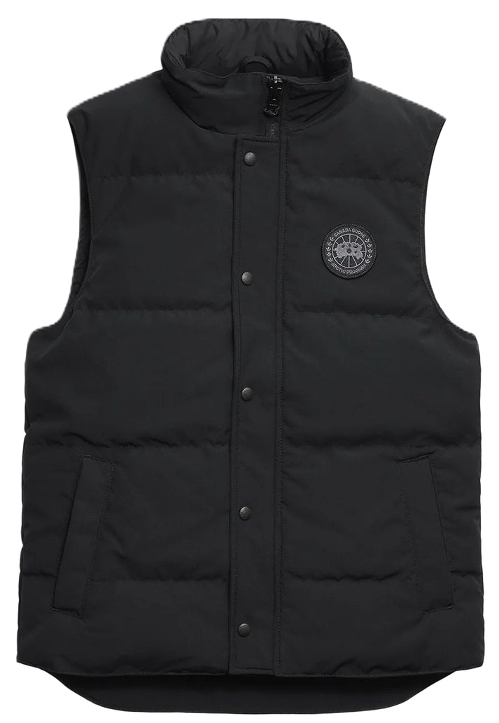 A black quilted puffer vest with a high collar, a central snap-button placket, two side pockets, and a round black and grey circular patch on the left chest.