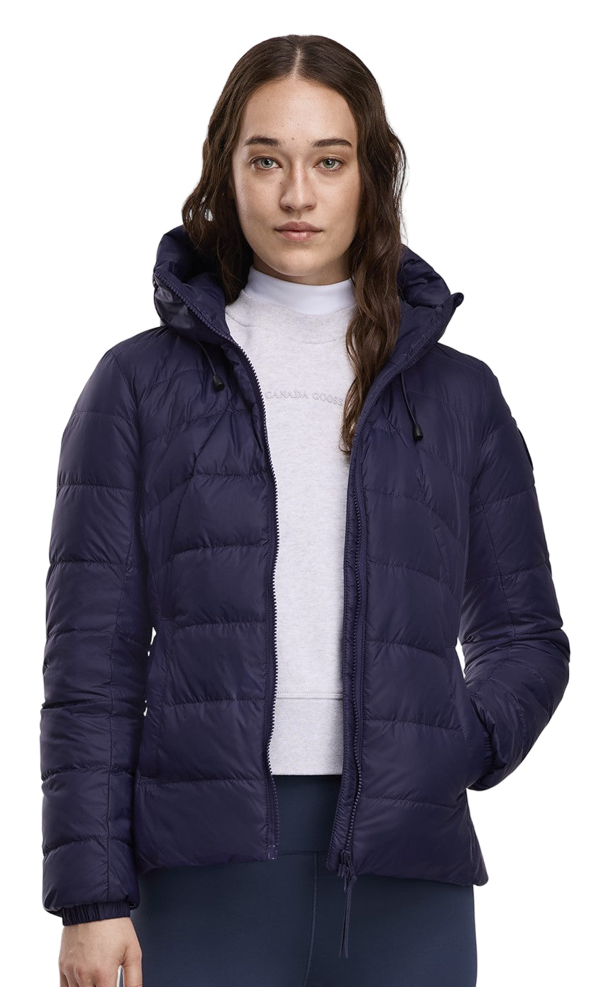 A woman with long brown hair wearing a dark blue puffer jacket and white turtleneck.