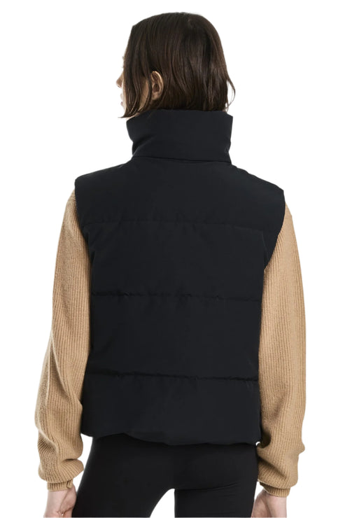 A person with short hair is facing away from the camera wearing a black puffy vest with a tall collar over a tan-colored sweater.