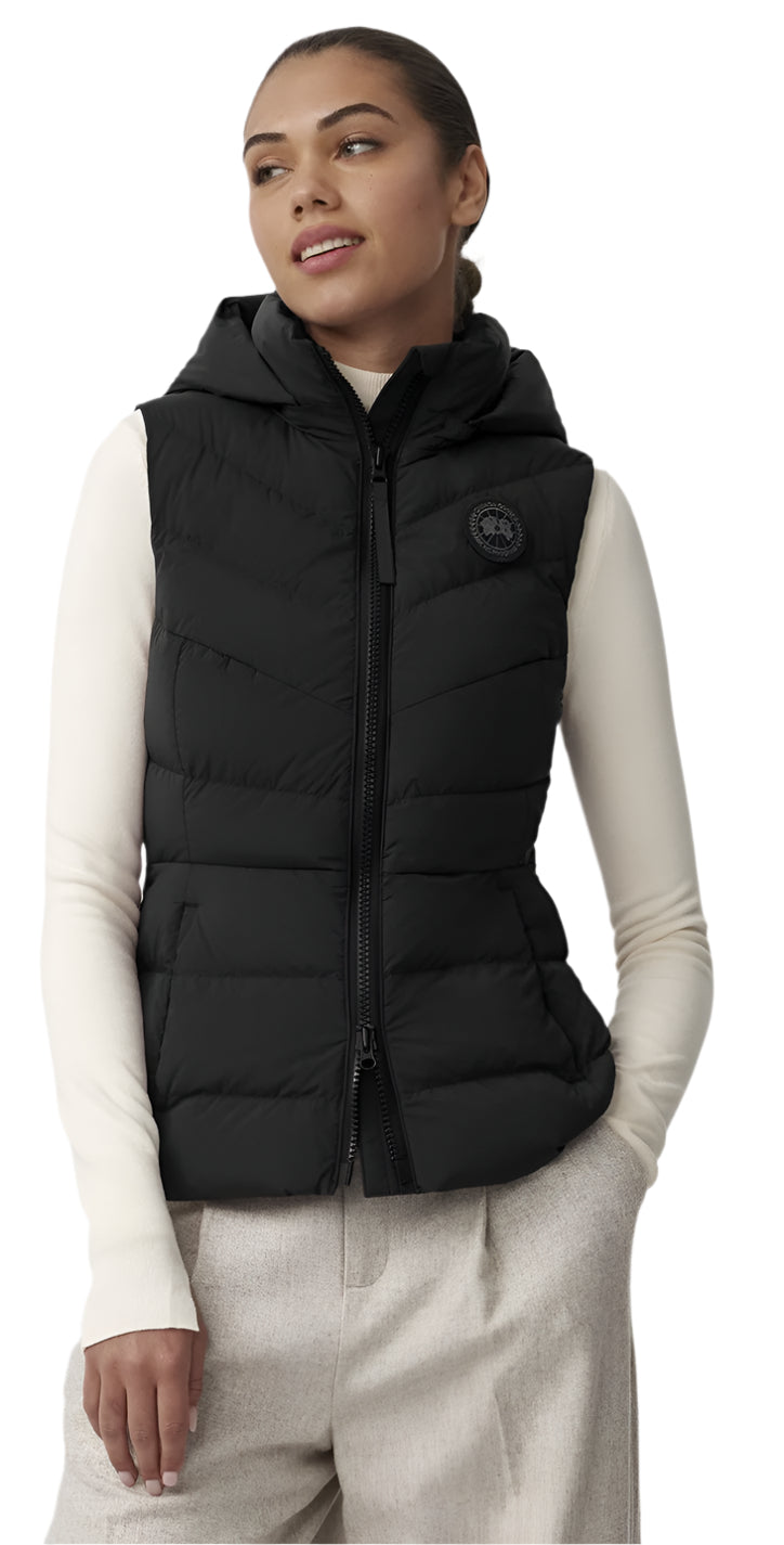 A woman wearing a black puffer vest with a hood and full zipper, with a circular logo on the upper right, and a long-sleeved cream shirt and white pants.