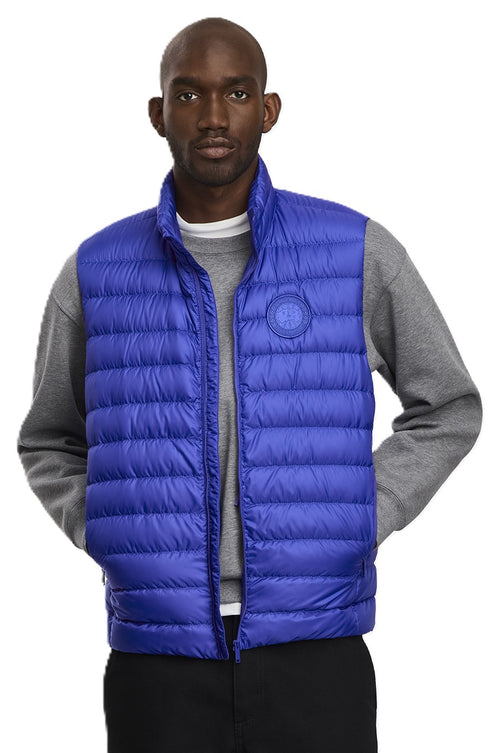 A dark-skinned person wearing a blue quilted vest with a round logo on the chest over a gray sweater, and black pants, looking at the camera.