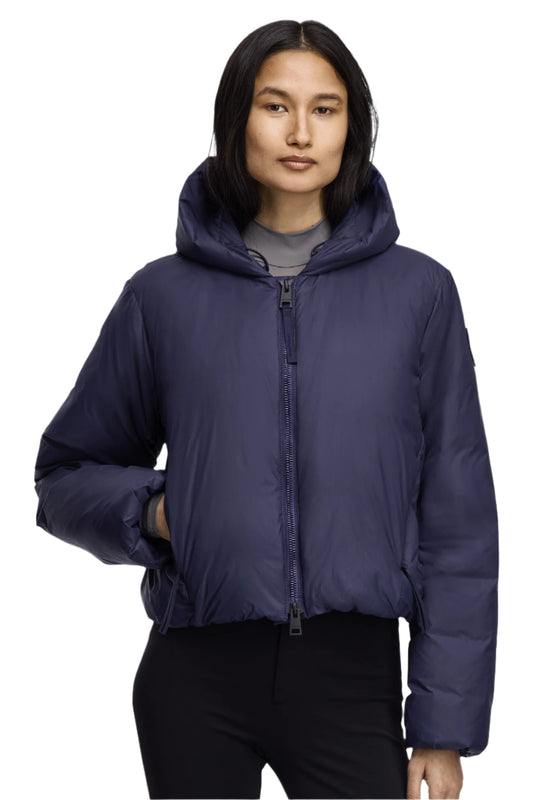 A woman in a short navy blue puffer jacket with a hood and a zipper, standing with one hand in her pocket.