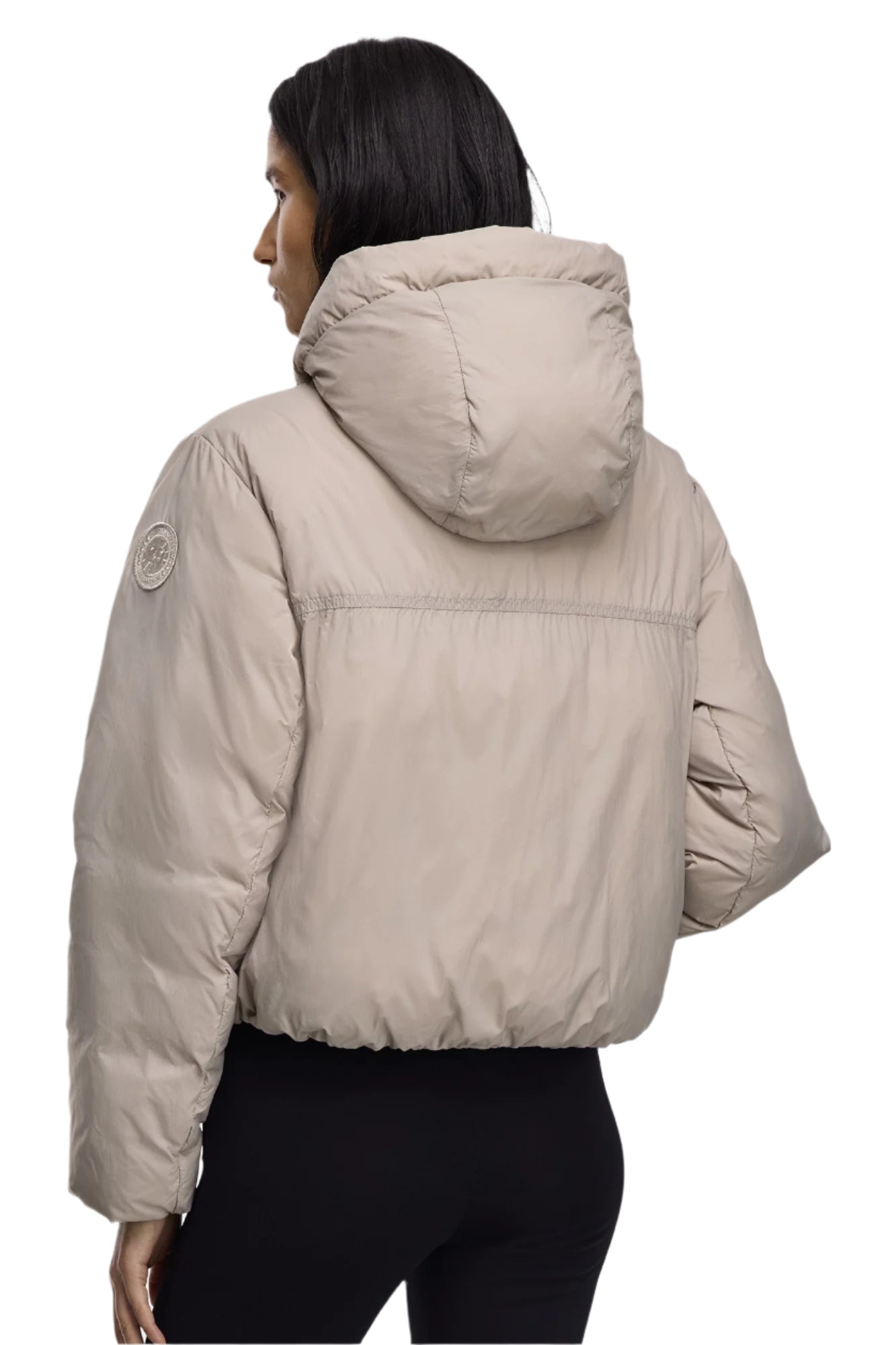 A woman stands facing left wearing a beige puffer jacket with a hood.