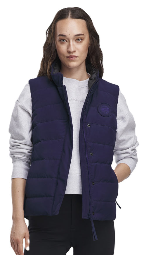 A woman wears a navy blue quilted puffer vest over a white sweatshirt. The vest has a round logo patch on the chest and an adjustable drawstring at the hem.