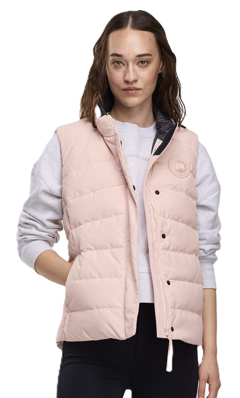 A woman wearing a light pink, quilted vest with black buttons and a white string. She wears a light grey sweatshirt under the vest.