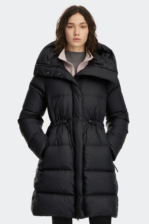 A person wearing a long, black, puffy winter coat with a large collar, with drawstrings cinching the waist.
