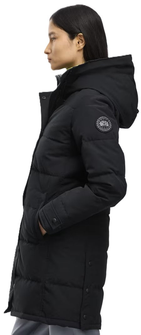 A woman seen from the side profile wearing a black hooded, mid-length puffer coat with a circular logo patch on the left upper sleeve.