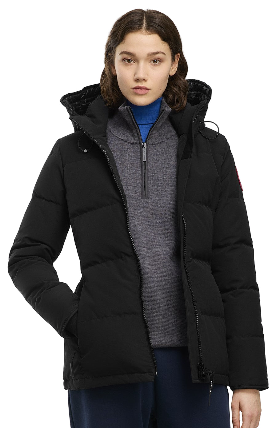 A woman wearing a black, hooded down winter jacket and a grey turtleneck, with her hand in her pocket.