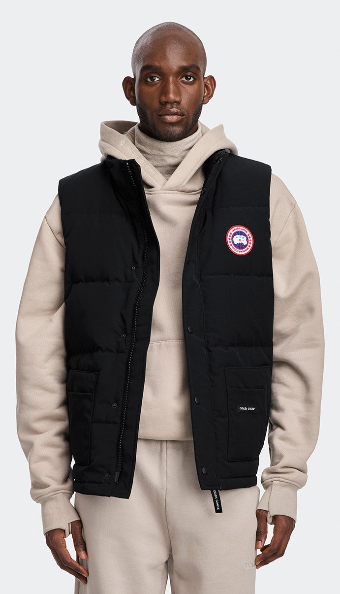 A man wearing a black puffy vest over a light-colored turtleneck and hoodie with matching sweatpants. The vest has the logo on the chest.