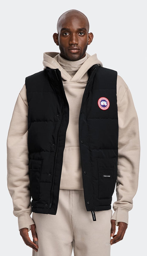 A man wearing a black puffy vest over a light-colored turtleneck and hoodie with matching sweatpants. The vest has the logo on the chest.