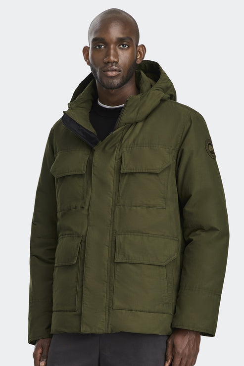A dark-skinned man wears an olive-green hooded parka jacket with many pockets.