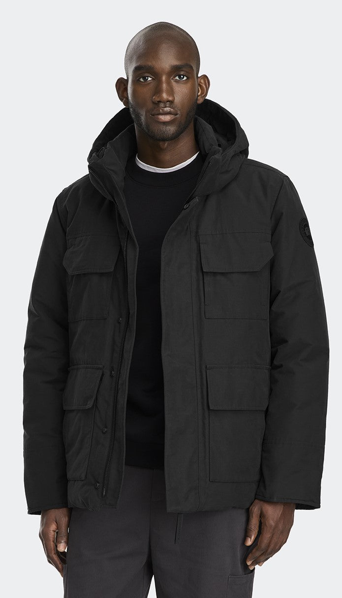 A Black man wearing a black hooded jacket with multiple front pockets and gray trousers.