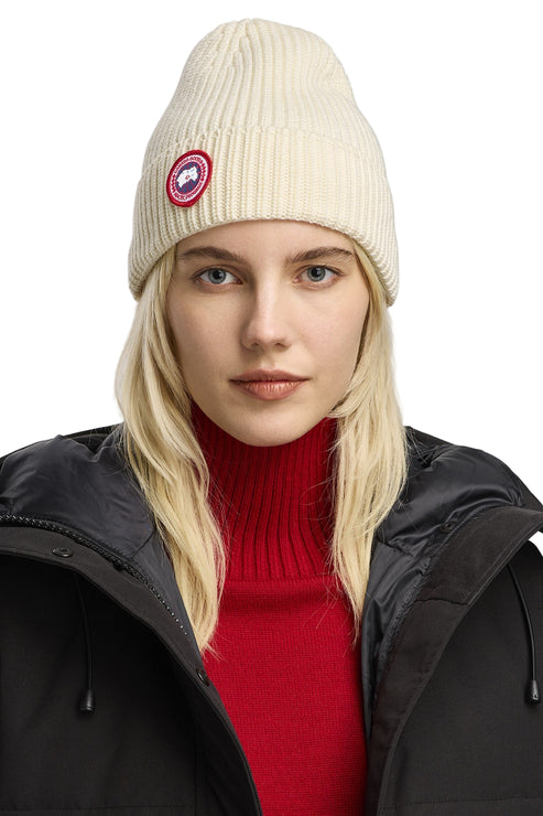 Person with blonde hair wearing a black coat, red turtleneck, and white ribbed beanie with a round logo patch.