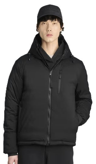 A person wearing a black, hooded, insulated puffer jacket with a full-length front zipper, a chest pocket with a vertical zipper, and a black baseball cap.