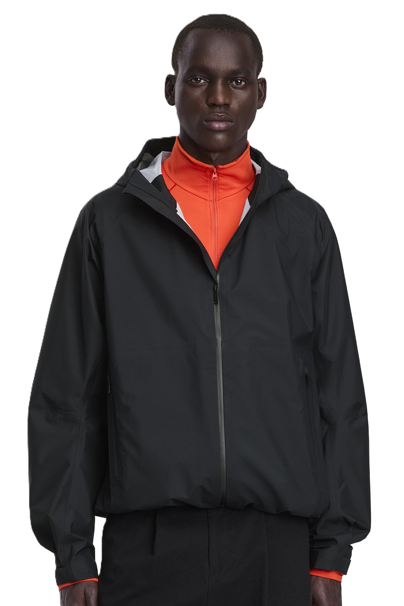 A Black man wears a black, hooded zip-up jacket over a red shirt.