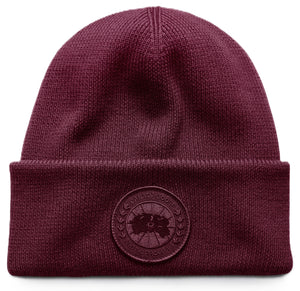 A deep burgundy ribbed knit beanie with a folded cuff featuring a circular tonal logo patch embroidered with text reading CANADA GOOSE ARCTIC PROGRAM.