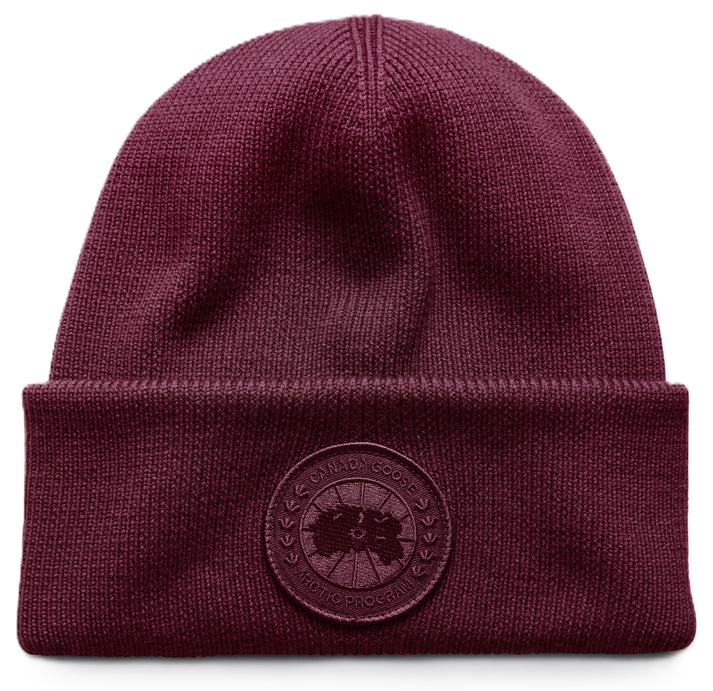 A deep burgundy ribbed knit beanie with a folded cuff featuring a circular tonal logo patch embroidered with text reading CANADA GOOSE ARCTIC PROGRAM.