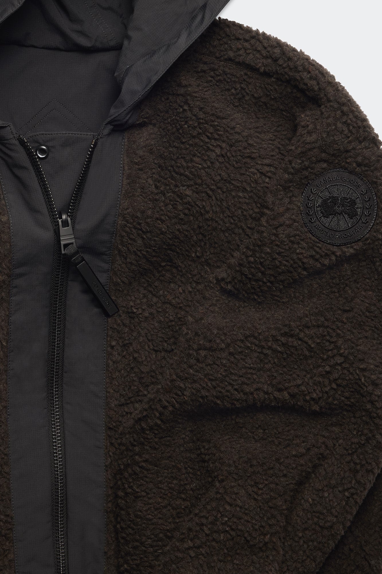 Close-up of a dark brown, fuzzy jacket with a black zipper and hood, and a black circular patch on the sleeve.