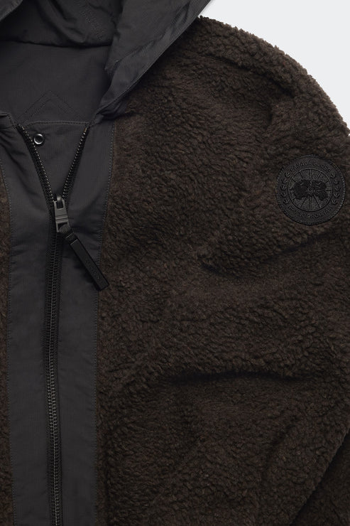 Close-up of a dark brown, fuzzy jacket with a black zipper and hood, and a black circular patch on the sleeve.