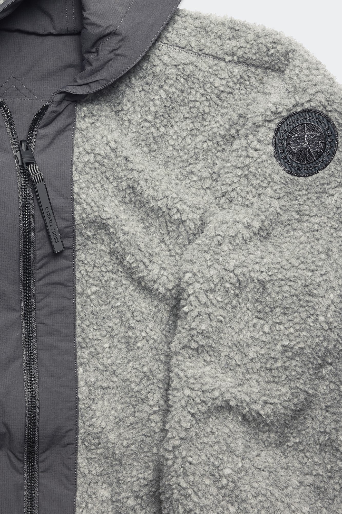 Close-up of a grey, teddy-fleece jacket with a black emblem patch on the right side and a grey, padded front with a visible zipper.