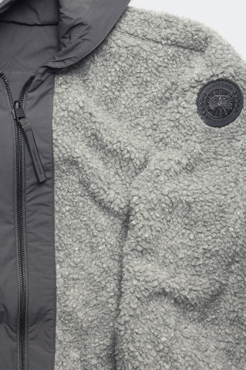Close-up of a grey, teddy-fleece jacket with a black emblem patch on the right side and a grey, padded front with a visible zipper.