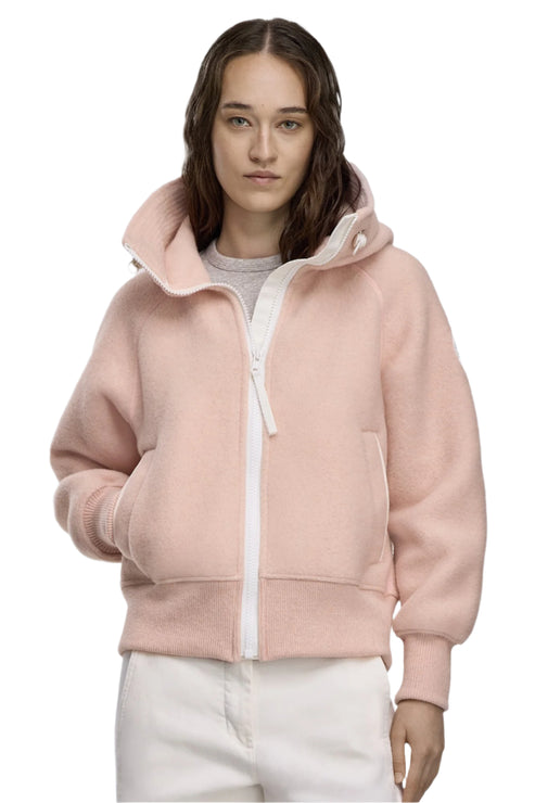 A person wearing a pink, hooded, zip-up jacket and white pants, with their hands in the jacket pockets.