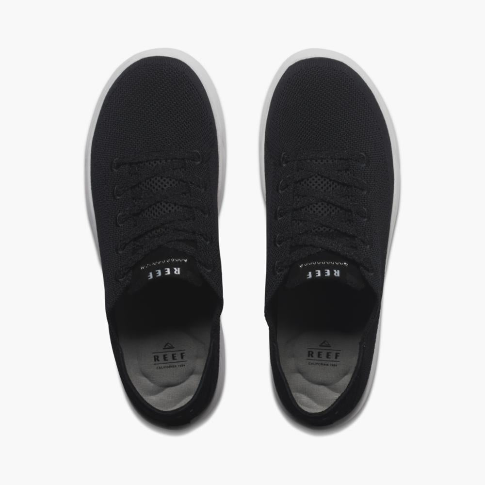 A pair of black sneakers with white soles, seen from above, showing shoelaces and brand labels on the inner sole.