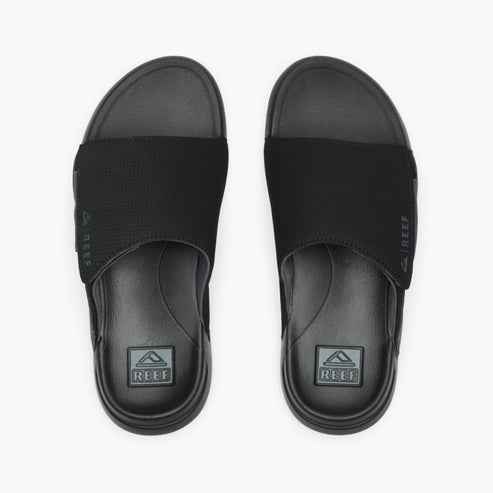 A pair of black slide sandals with fabric strap and logo on the footbed.